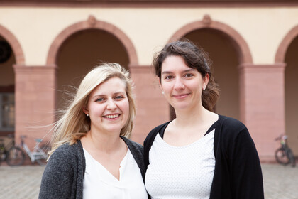Zwei als Frauen gelesene Personen stehen lächelnd nebeneinander vor den Bogengängen des Ehrenhof West. Die Person links hat blondes Haar und trägt einen grauen Cardigan über einer weißen Bluse, die Person rechts hat braunes Haar, trägt einen schwarzen Cardigan über einem weißen Top mit Punkten.