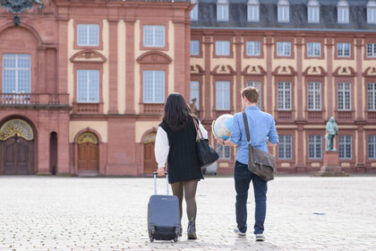 Hintergrund: Schloss Mannheim auf dem Ehrenhof, links läuft eine Frau, die einen Koffer hinter sich her zieht und rechts neben ihr läuft ein Mann mit einen Globus in der Hand
