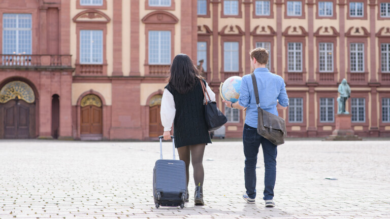 Hintergrund: Schloss Mannheim auf dem Ehrenhof, links läuft eine Frau, die einen Koffer hinter sich her zieht und rechts neben ihr läuft ein Mann mit einen Globus in der Hand