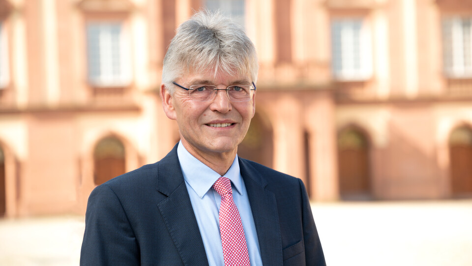 Prof. Spengel hat kurze graue Haare und trägt Anzug und Krawatte. Im Hintergrund ist das Schloss zu sehen.
