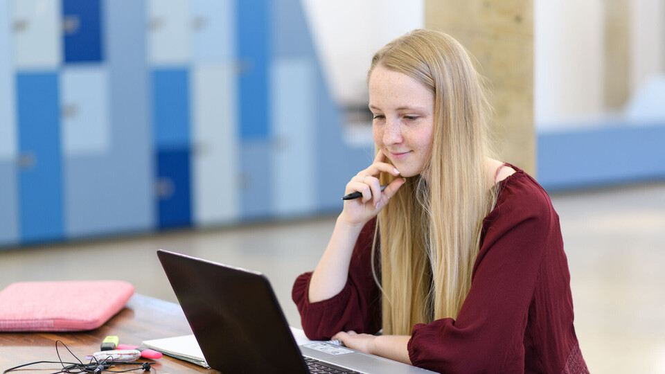 Online-Lehre in der Universität Eine Studentin sitzt mit ihrem Laptop an einem Tisch und betrachtet etwas auf dem Bilschirm.
