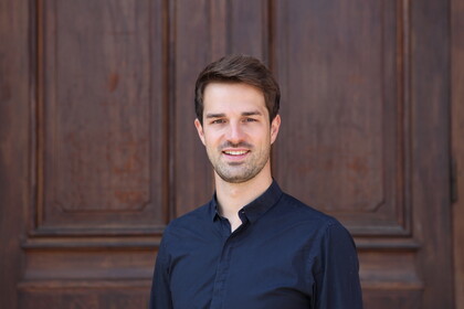 Portrait von Valentin Lang. Er trägt ein dunkelblaues Hemd und steht lächelnd vor einer Holztüre.