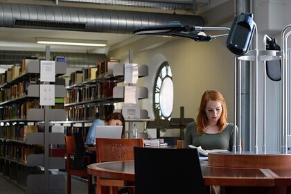 Zwei Studierende sitzen in der Bibliothek. Vorne rechts sitzt eine Studierende, die rote Haare hat und liest ein Buch. Hinter ihr sitzt eine Studierende mit braunen Haaren vor ihrem Laptop.
