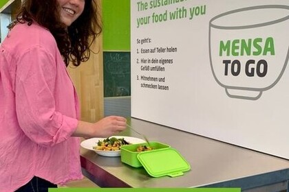 Weiblich gelesene Studierende steht an einem Tisch in der Mensa und füllt Essen von einem Teller in eine grüne Plastikbox. Vor ihr an der Wand hängt ein Schild, das das "Mensa to go"-System bewirbt. Sie trägt ein pinkes Oberteil und schaut lächelnd über ihre rechte Schulter in die Kamera.