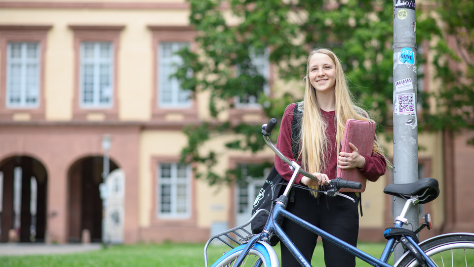 Studentin mit blauem Fahrrad vor sich vor dem Schloss