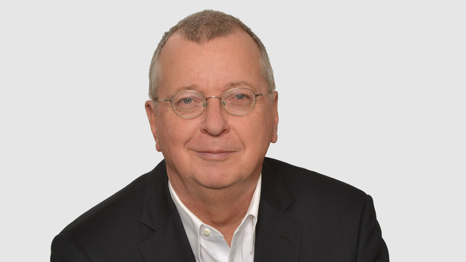 Prof. Weber hat kurze graue Haare und eine Brille. Er trägt ein Jacket und ein weißes Hemd.