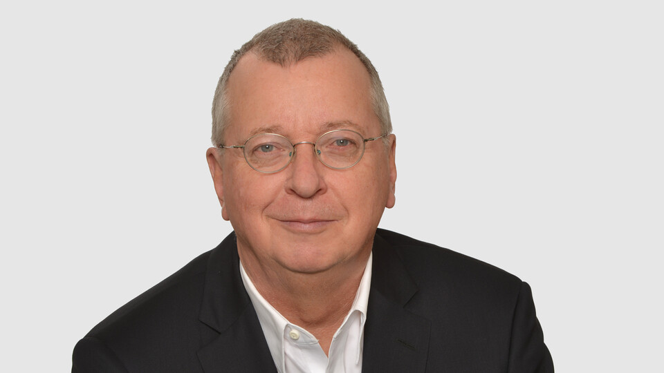 Prof. Weber has short gray hair and glasses. He is wearing a jacket and a white shirt.