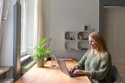 Person mit langen blonden Haaren und grünem Pulli sitzt mit Laptop an einem Schreibtisch am Fenster
