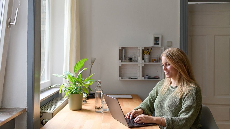 Person mit langen blonden Haaren und grünem Pulli sitzt mit Laptop an einem Schreibtisch am Fenster