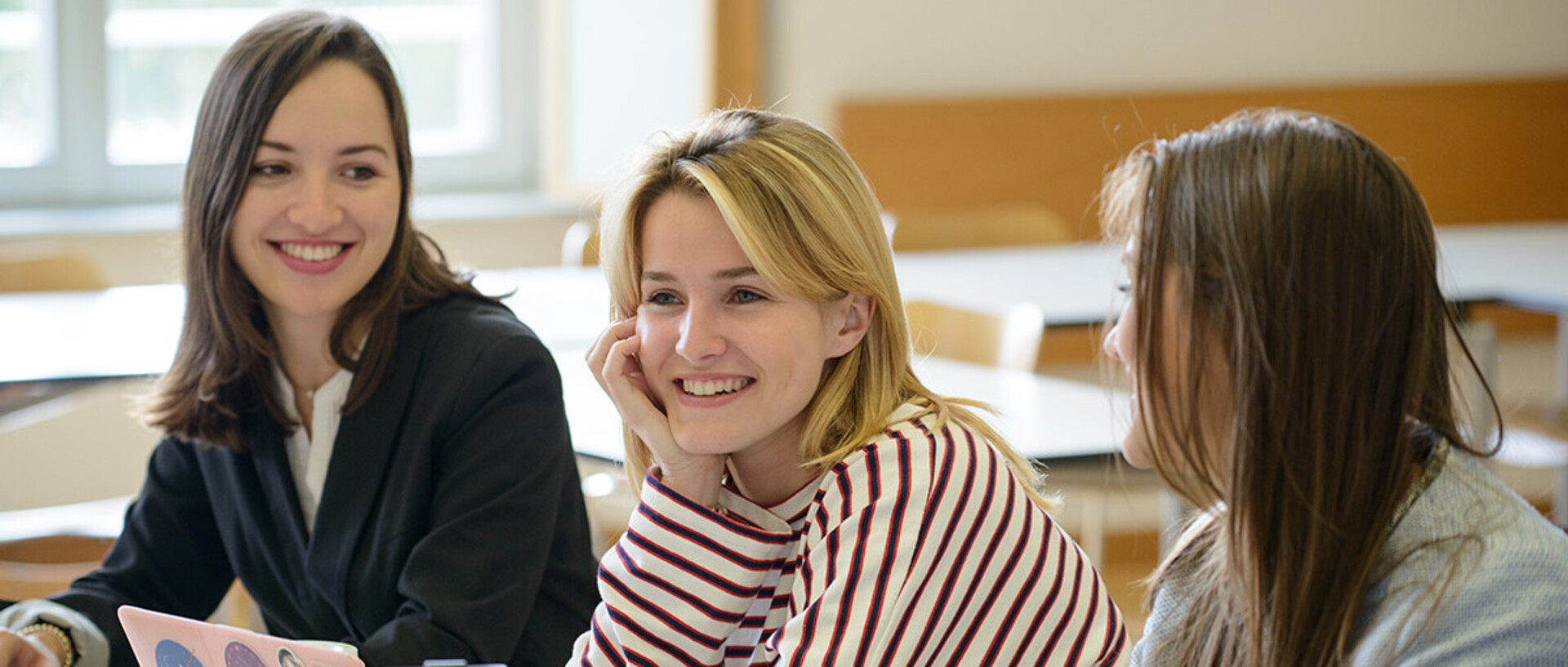 Drei Studentinnen sitzen lachend in einem Seminarraum.