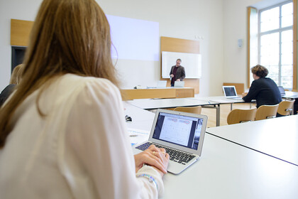 Studierende in einem Seminarraum, am Laptop, eine Lehrperson steht an der Tafel und erklärt