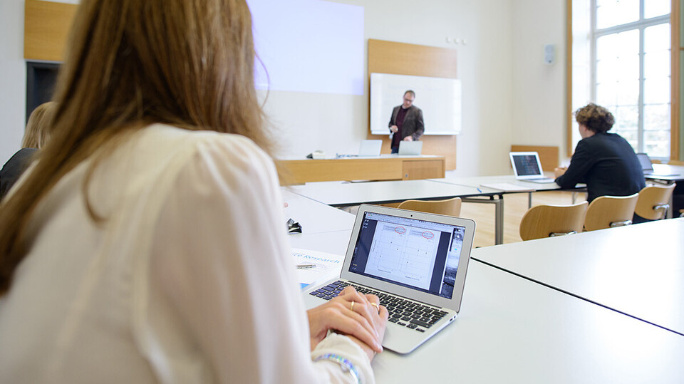 Studierende in einem Seminarraum, am Laptop, eine Lehrperson steht an der Tafel und erklärt