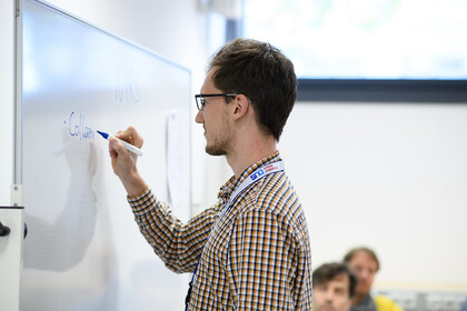 Ein Dozent schreibt mit blauem Stift auf ein Whiteboard, während im Hintergrund zwei Personen zum Dozenten blicken.