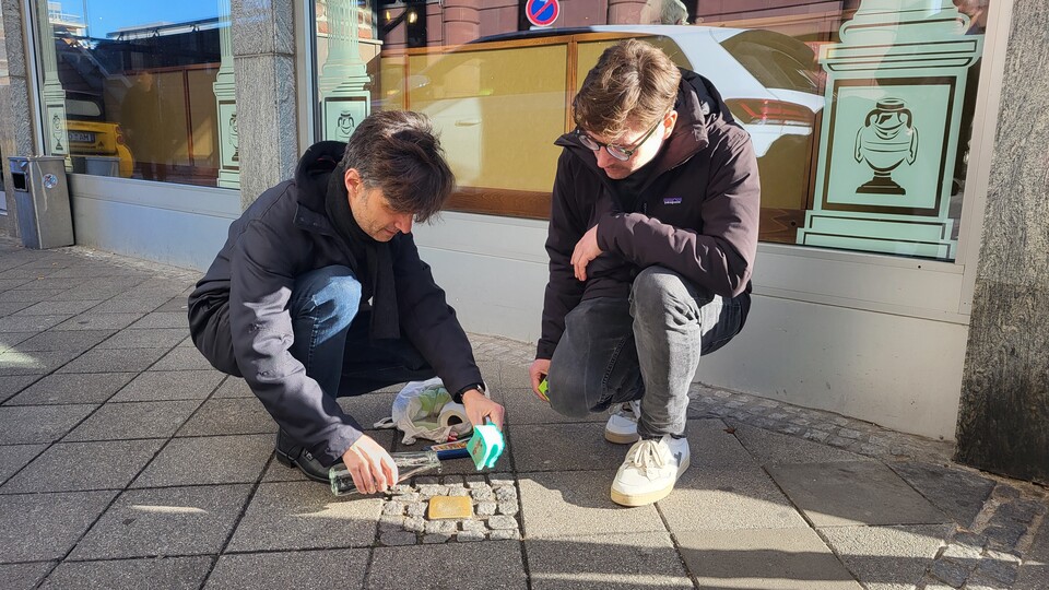 Zwei Mitglieder des Otto-Selz-Instituts, links Professor Alpers und rechts Sven Cornelisse, knien auf dem Gehweg neben dem Stolperstein von Otto Selz, den sie gerade reinigen. Professor Alpers hält eine Flasche und einen Putzlappen, während Sven Cornelisse aufmerksam zusieht. Im Hintergrund spiegelt sich die urbane Umgebung in den Fensterscheiben eines Gebäudes.