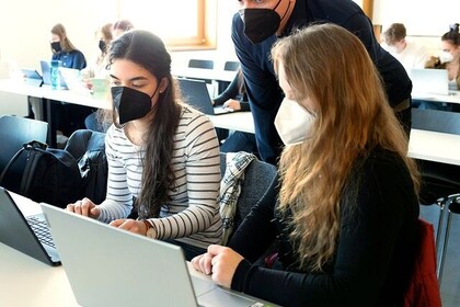 Zwei weiblich gelesene Studierende sitzen in einem Seminarraum vor Laptops. Eine männlich gelesene dozierende Person blickt ihnen über die Schulter. Alle tragen FFP-2-Masken, im Hintergrund sitzen weitere Studierende.