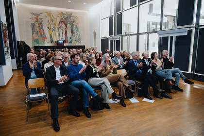 Viele Personen sitzen in einem großen Raum auf Stühlen und klatschen.