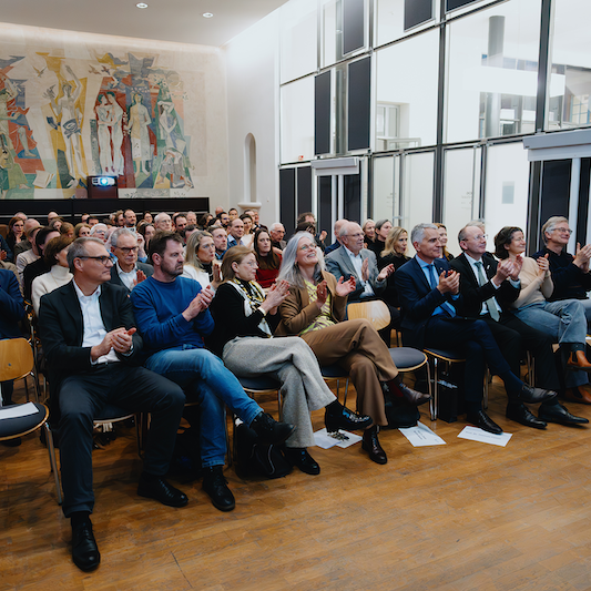 Viele Personen sitzen in einem großen Raum auf Stühlen und klatschen.