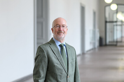 Portrait von Jochen Taupitz. Er trägt einen grünen Anzug und steht in einem Flur.