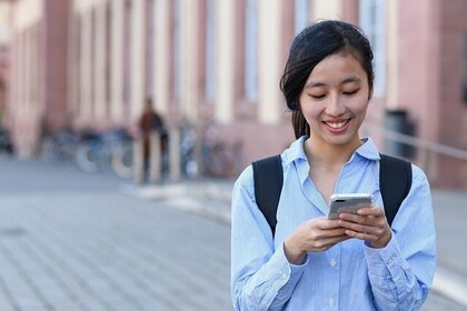 Eine weiblich gelesene Studierende blickt lächelnd auf ihr Smartphone. Schräg hinter ihr ist das Hauptgebäude der Universität zu sehen.