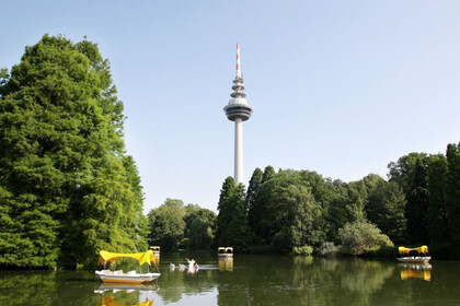 Auf dem Luisenparksee fahren kleine Boote mit gelben Dächern. Die Sonne scheint und der Himmel ist blau. Das Ufer ist gesäumt von großen Bäumen mit dichten, grünen Baumkronen. Im Hintergrund ragt der Fernmeldeturm empor. Link: Instagram-Post Jugendgruppe des badischen Blinden- und Sehbehindertenvereins