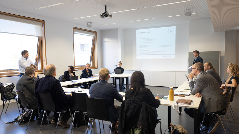Eine Gruppe von Menschen sitzt an Tischen in einem Seminarraum. An die Wand wird eine Präsentation projiziert.