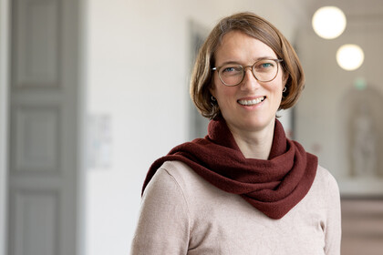 Astrid Lembke trägt einen beige-farbenen Pullover sowie einen roten Schal und steht in einem Flur. Die Person heißt Astrid Lembke. 