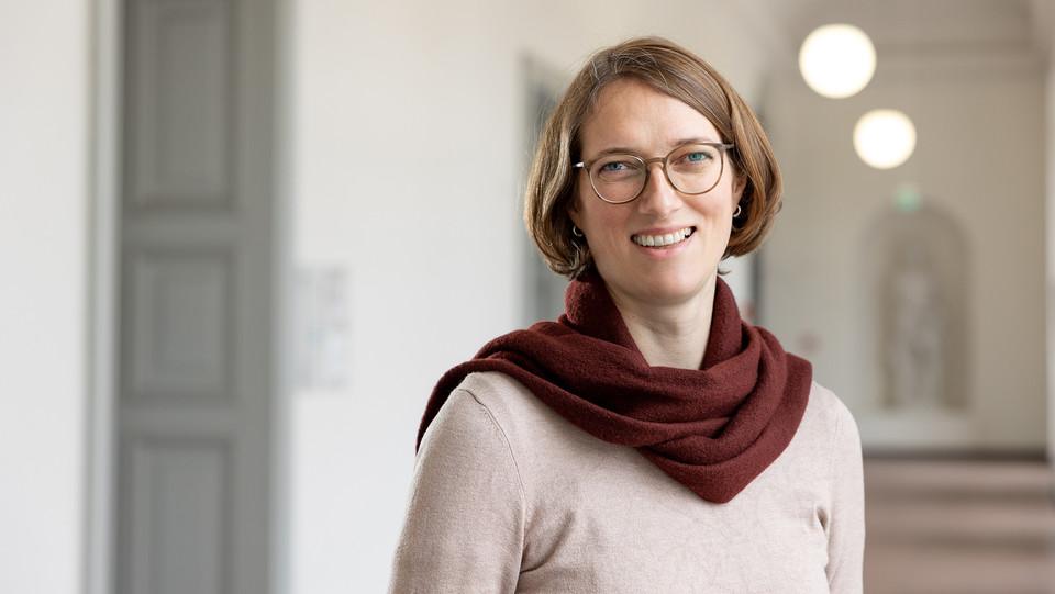 Astrid Lembke trägt einen beige-farbenen Pullover sowie einen roten Schal und steht in einem Flur. Die Person heißt Astrid Lembke. 