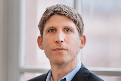 Eine Person trägt ein hellblaues Hemd sowie einen schwarzen Blazer und steht vor einem Fenster. Die Person heißt Torsten Biemann.