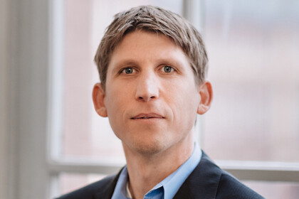 Eine Person trägt ein hellblaues Hemd sowie einen schwarzen Blazer und steht vor einem Fenster. Die Person heißt Torsten Biemann.