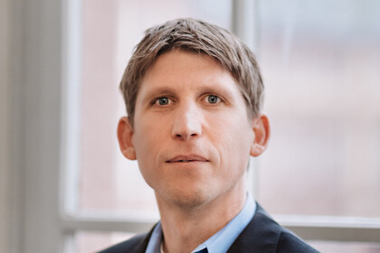 Eine Person trägt ein hellblaues Hemd sowie einen schwarzen Blazer und steht vor einem Fenster. Die Person heißt Torsten Biemann.