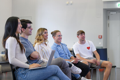 Fünf Studierende auf Stühlen in einer Reihe, lachend und nach vorne blickend