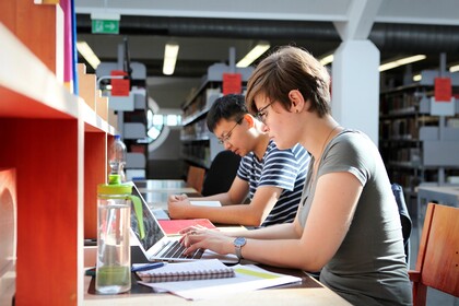 Eine Studentin und ein Student sitzen in der Bibliothek nebeneinander und lernen.