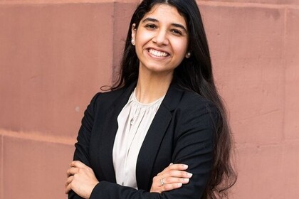 Porträt der Studentin Tushna aus Indien. Sie trägt einen schwarzen Blazer und eine weiße Bluse und hat die Arme vor der Brust gekreuzt. Ihr langes schwarzes Haar trät sie offen. Sie lacht freundlich in die Kamera. Link: Instagram Post myUniMa
