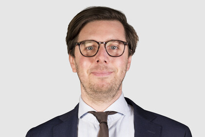 Eine Person trägt ein hellblau-weiß gestreiftes Hemd, eine braune Krawatte sowie einen dunkelblauen Blazer und steht vor einer weißen Wand. Die Person heißt Philipp Dörrenberg.