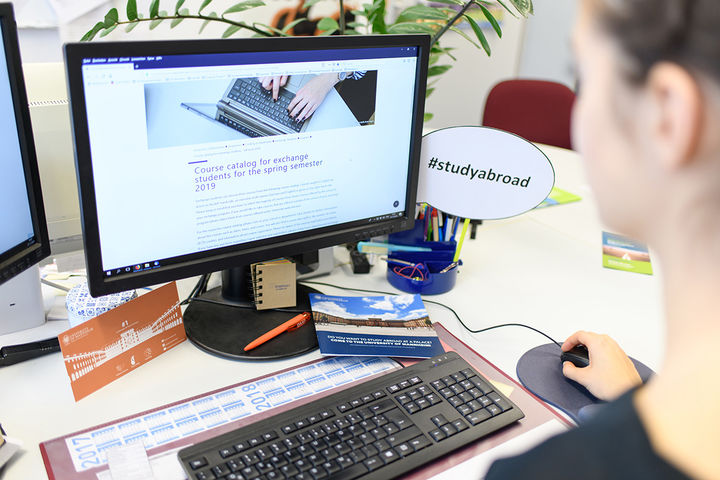 A person is sitting at a desk and using a computer. The computer screen shows a university website with the title "Course catalog for exchange students for the spring semester 2019". There is a sign on the computer that says "#studyabroad". 