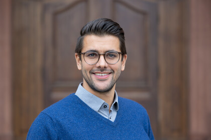 Lukas Hack. He is wearing a blue sweater and is standing in front of a brown door.