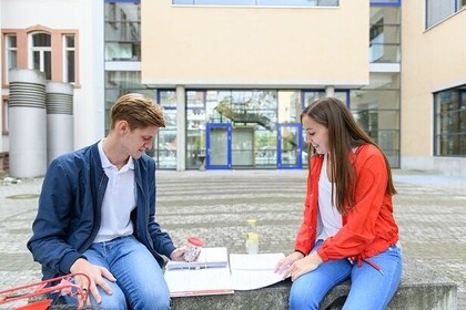 Zwei Studierende sitzen auf dem Uni-Gelände. Der Student links hat kurze hellbraune Haare und trägt ein weißes Oberteil mit blauer Jacke. Die Studentin links hat lange braune Haare und trägt ein weißes Oberteil mit roter Jacke. Beide schauen auf ihre Unterlagen, die vor ihnen liegen.