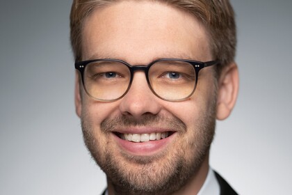 Eine Person trägt ein weißes Hemd sowie einen schwarzen Blazer und steht vor einer grauen Wand. Die Person heißt Marc Lerchenmüller.