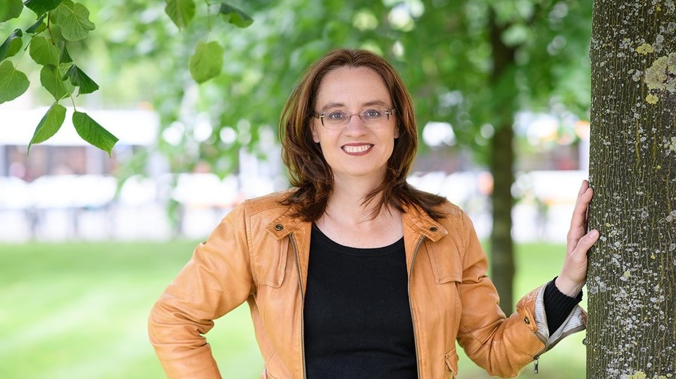 Portrait von Michele Tertilt. Sie trägt einen schwarzen Pollover sowie eine orange farbige Jacke und steht auf einer Grünfäche.