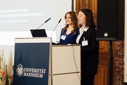 Zwei Personen stehen hinter einem Rednerpult. Die Person links hat lange hellbraune Haare und trägt eine blaue Bluse. Die Person rechts hat lange dunkelbraune Haare und trägt ein weißes Hemd mit einem schwarzen Blazer.