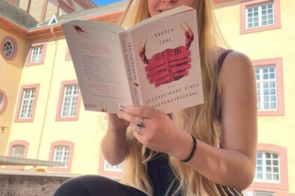 Eine Studentin sitzt im Schlosshof und liest das Buch "Beschreibung einer Krabbenwanderung" von Karosch Taha. Link: Instagram-Post Mannheim liest ein Buch