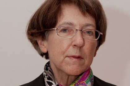 Portrait von Ursula Wolf. Sie trägt einen bunten Schal sowie einen schwarzen Blazer und steht vor einer grauen Wand.