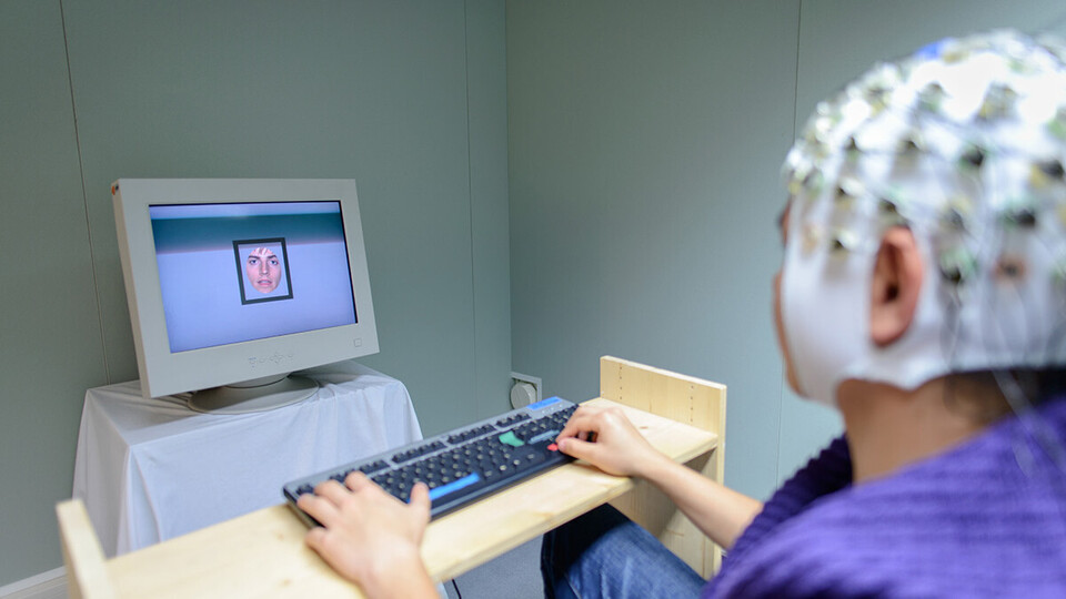 Eine Person trägt eine Haube mit Elektroden auf dem Kopf und sitzt an einer Tastatur vor einem Monitor.