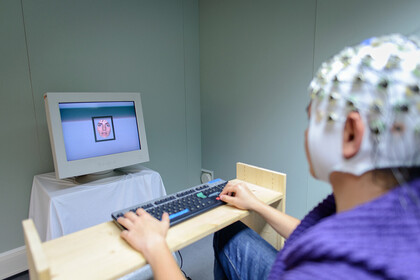 Eine Person trägt eine Haube mit Elektroden auf dem Kopf und sitzt an einer Tastatur vor einem Monitor.