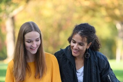Zwei Studierende sitzen auf einer Wiese und schauen lächelnd in ein Heft. Im Hintergrund verschwommene Bäume.