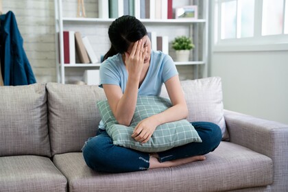 Eine Frau sitzt im schneidersitz auf dem Sofa. Auf ihrem Schoß liegt ein Kissen, sie fasst sich an die Stirn. 