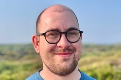 Christopher Klamm vor einer Wiese und blauem Himmel im Sonnenschein. Er trägt eine Brille und einen blauen Pullover.