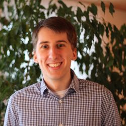 Portrait photo of a smiling man with brown hair and blue plaid shirt in front of a background of plants.  