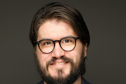 Rainer Freudentaler vor einem dunklen Hintergrund. Er trägt eine Brille, ein dunkelrotes Shirt und darüber einen karierten Blazer.