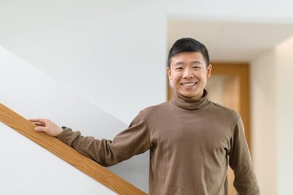 Jianming Cui aus Kanton. Er trägt einen braunen Rollkragenpullover und legt seinen rechten Arm lässig auf ein Treppengeländer.